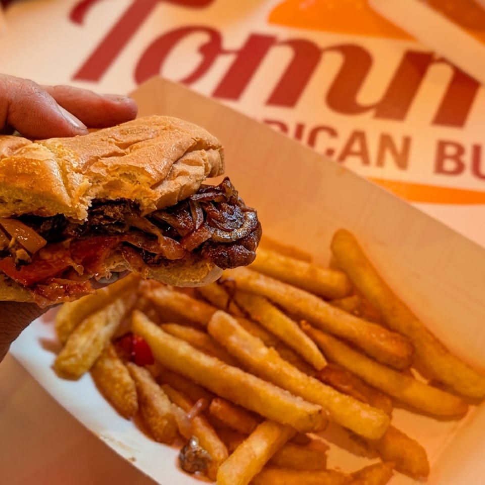 A person's hand holds a loaded sandwich over a tray of golden fries at Tommy's.
