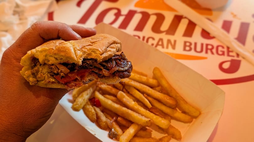 A person's hand holds a loaded sandwich over a tray of golden fries at Tommy's.