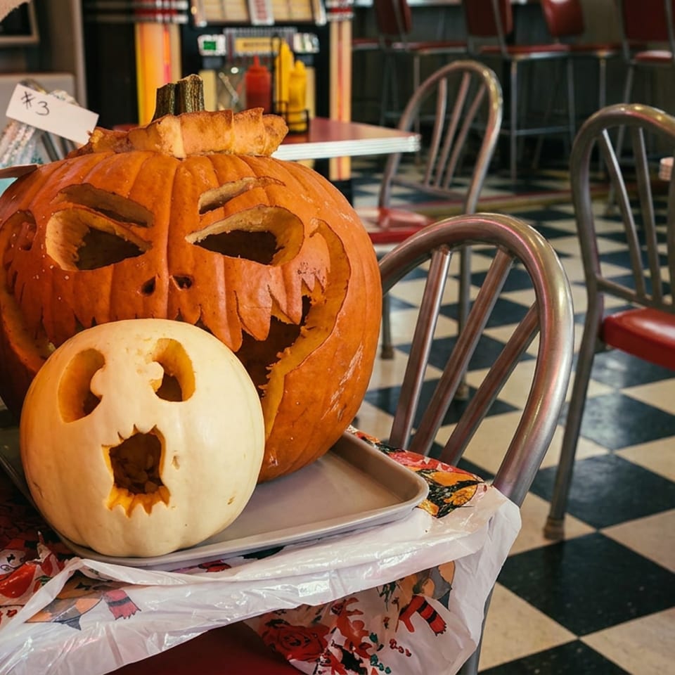 Jack-o'-lanterns and a white carved pumpkin on a chair in a diner with checkered floor.