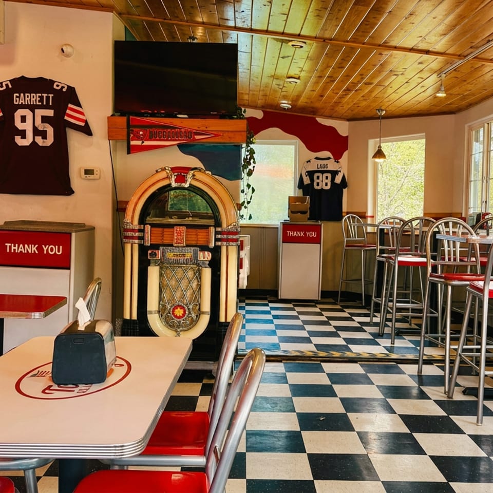 Retro diner interior with a jukebox, checkered floor, red seating, and sports jerseys on the walls.