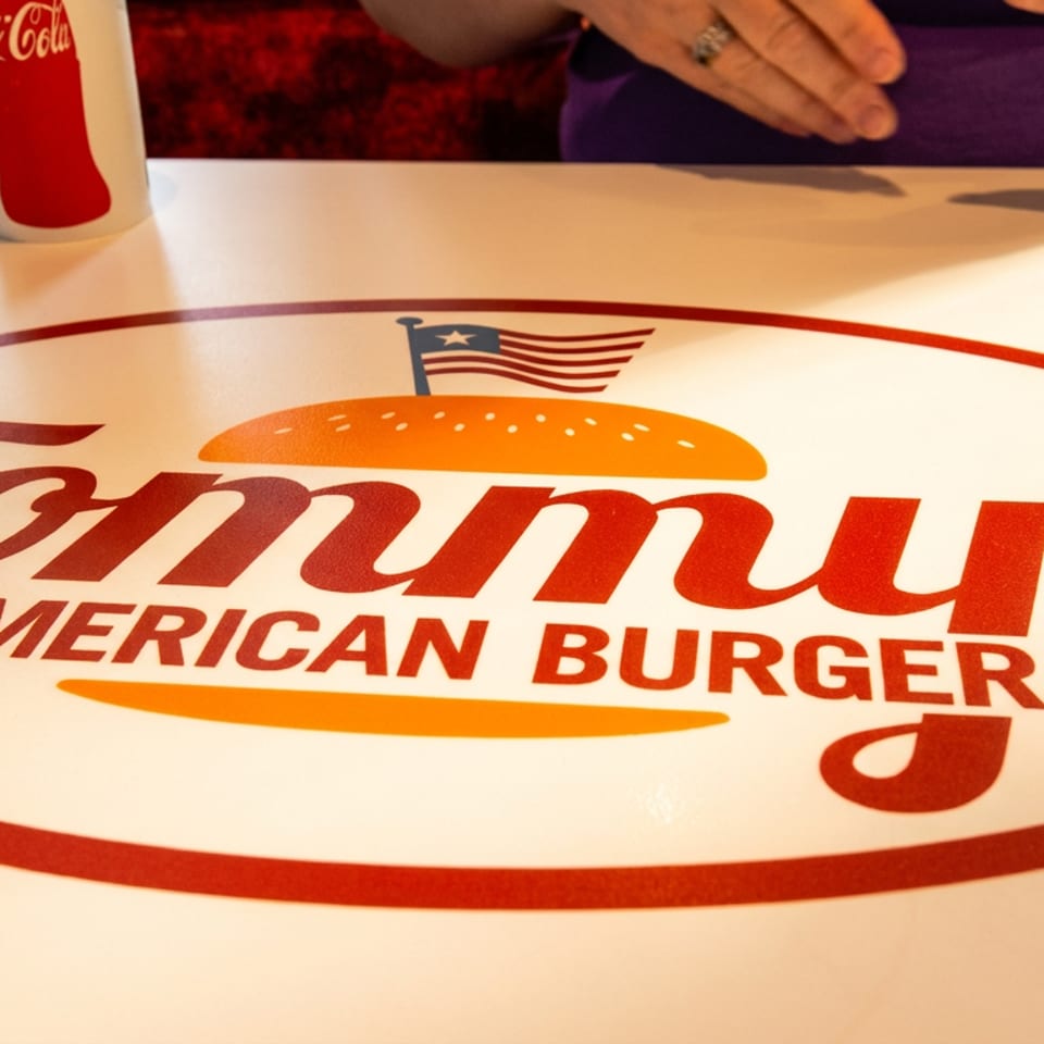 Tommy's All-American Burger Co. logo on a white table with a Coca-Cola cup and cutlery.