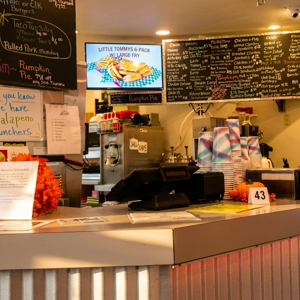 Busy restaurant counter featuring chalkboards, a digital menu, POS system, and stacked paper cups.