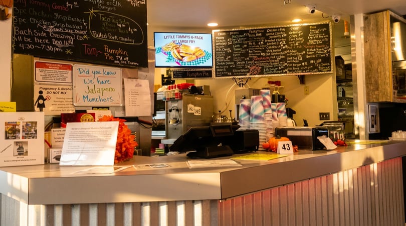 Busy restaurant counter featuring chalkboards, a digital menu, POS system, and stacked paper cups.